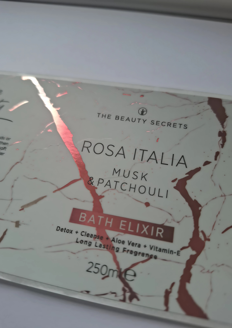 A close-up of a beauty product label for "The Beauty Secrets Rosa Italia Musk & Patchouli Bath Elixir" on a marble design background with rose gold accents.