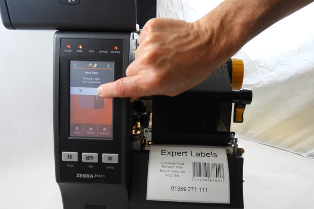 A close-up, eye-level shot shows a person's hand pressing a touchscreen on a Zebra ZT411 industrial printer. The printer's screen displays a message that reads, "Head Open: Printhead is open. Close the printhead." A label with a barcode and the text "Expert Labels" is visible on the printer's output tray.